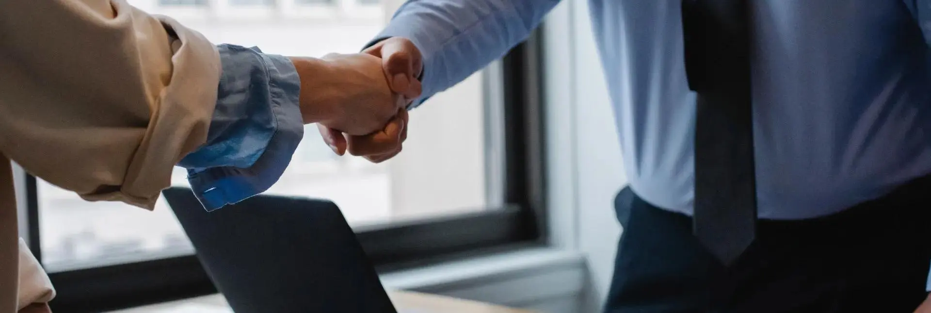 A close-up of a professional business handshake between a global procurement manager and a plastic packaging supplier in an office setting with a laptop.